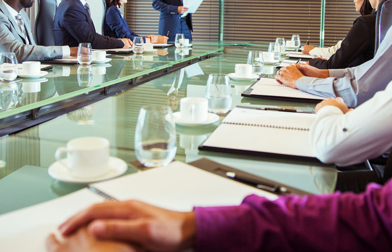 Image of business people sitting around a  boardroom glass table with note books, glasses of water and cups on the table. 