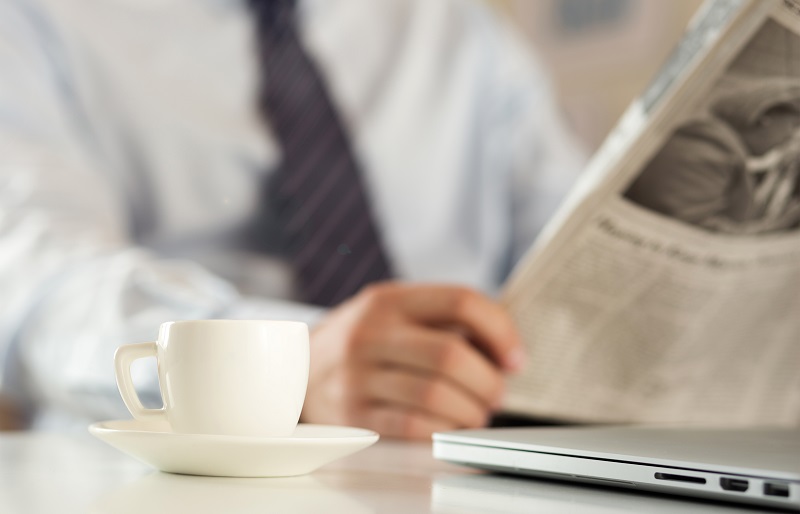 Man in business attire reading a newspaper beside a laptop and white cup and saucer
